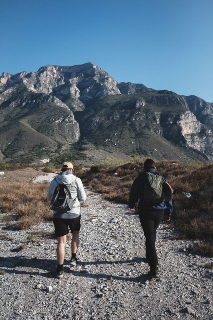 Two men backpack toward a mountain range under a clear sky. 